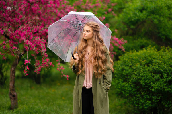 Pretty young blonde girl with long curly hair in vintage lace blouse and green cloak standing in spring park near pink blossom flowers. Tenderness romantic model posing with transparent umbrella.
