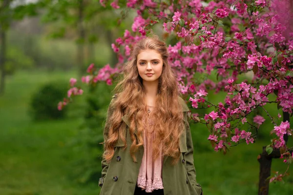 ピンクの花の近くの春の公園に立ってヴィンテージレースブラウスと緑のマントで長い巻き毛を持つかなり若いブロンドの女の子 優しさロマンチックなモデルポーズとカメラを見て — ストック写真