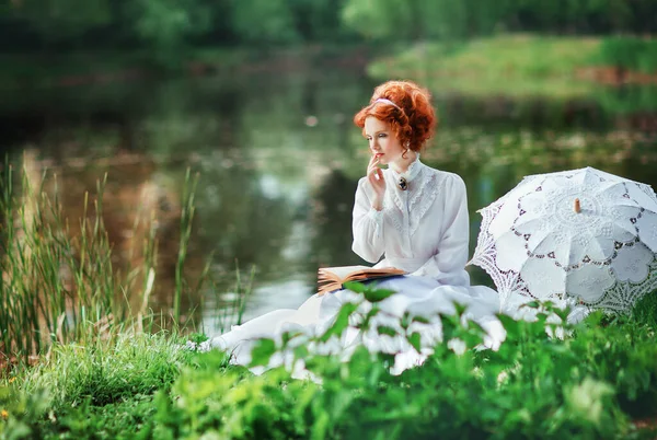 美丽的红头发姑娘 身穿白色长衫 坐在河边看书 带着花边伞的浪漫女人的艺术作品 — 图库照片