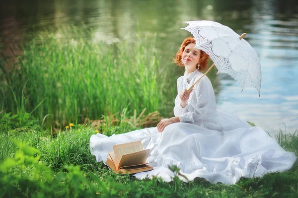 川の近くの芝生の上に座って白い長いヴィンテージのドレスで美しい赤髪の女の子 本やレースの傘とロマンチックな女性の芸術作品 かなりモデルの笑顔とカメラを見て — ストック写真