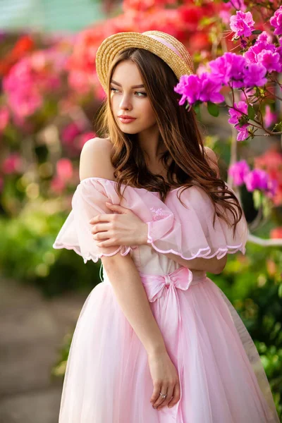美丽的女孩穿着粉红的古装 戴着草帽 站在五彩缤纷的花朵旁边 浪漫女人的艺术作品 漂亮温柔的模特 摆出姿势 看着镜头 — 图库照片