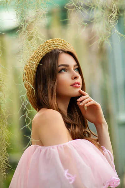 漂亮的女孩穿着粉红色的老式衣服 戴着草帽 站在绿色的背景上 种着植物 浪漫女人的艺术作品 漂亮温柔的模特 摆出姿势 看着镜头 — 图库照片