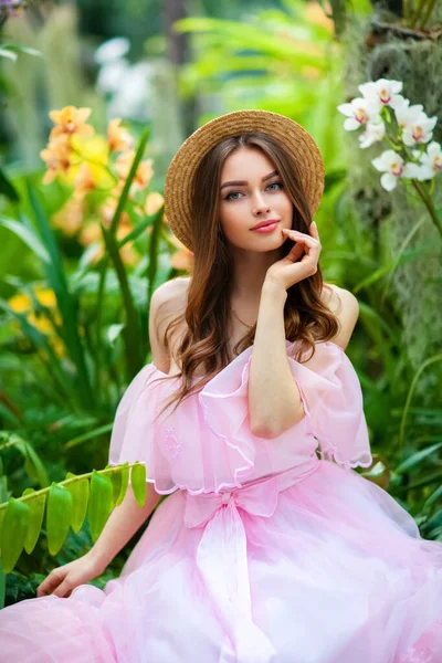 Renkli çiçeklerin yanında duran pembe klasik elbiseli ve hasır şapkalı güzel kız. Romantik bir kadının sanat eseri. Sevimli bir model, poz veriyor ve kameraya bakıyor..