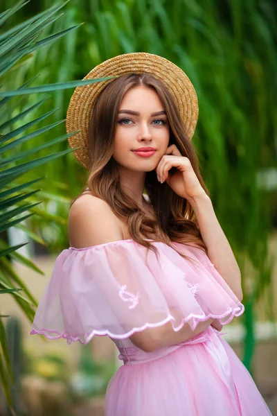 漂亮的女孩穿着粉红色的老式衣服 戴着草帽 站在绿色的背景上 种着植物 浪漫女人的艺术作品 漂亮温柔的模特 摆出姿势 看着镜头 — 图库照片