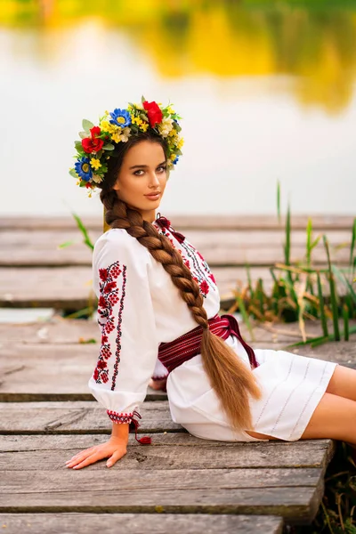 美丽的长长的辫子女 身穿乌克兰传统服饰 头戴花环 在河边的绿色自然中摆姿势 色彩艳丽 背景温暖的年轻貌美女子的画像 — 图库照片