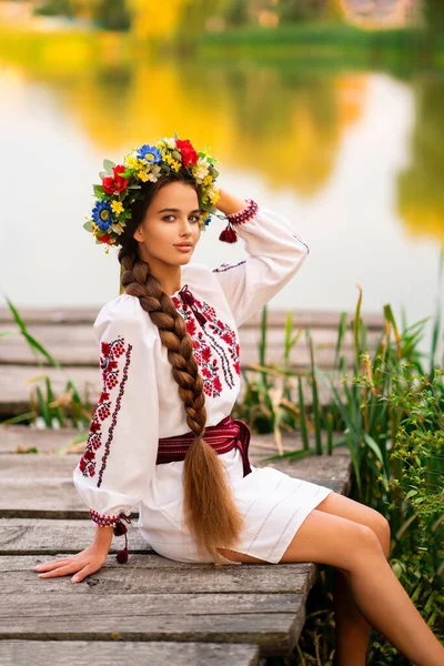 美丽的长长的辫子女 身穿乌克兰传统服饰 头戴花环 在河边的绿色自然中摆姿势 色彩艳丽 背景温暖的年轻貌美女子的画像 — 图库照片