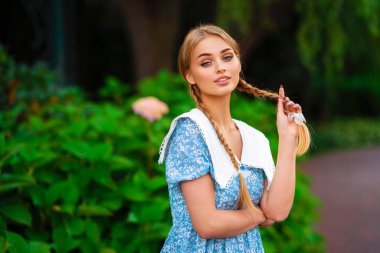 Kısa mavi elbiseli, güzel, genç, sarışın, gülümseyen bir kız, beyaz bisikletin yanında duruyor, elinde kocaman bir buket dolusu odunsu bir sepet var. Açık havanın tadını çıkaran bir bayan. Çiçekli yeşil arka plan..