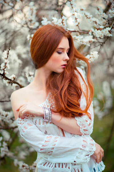Spring Beautiful romantic red haired girl in white lace dress standing in blooming garden. Dreaming young model looking down.