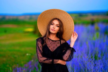 Renkli lavanta tarlasında duran siyah şeffaf elbiseli ve samandan büyük şapkalı güzel bir kızın portresi. Romantik bir kadının sanat eseri. .