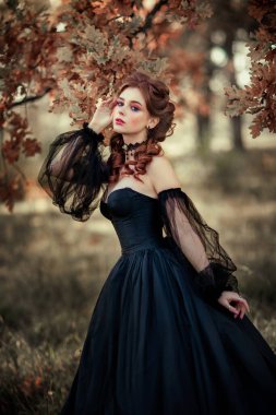 Sonbahar ağacının yanında duran muhteşem moda gotik kız portresi. Fantezi sanat çalışmaları. Siyah elbiseli ve şapkalı kızıl saçlı manken kamera ve pozlara bakıyor. 