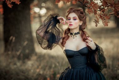 Sonbahar ağacının yanında duran muhteşem moda gotik kız portresi. Fantezi sanat çalışmaları. Siyah elbiseli ve şapkalı kızıl saçlı manken kamera ve pozlara bakıyor. 
