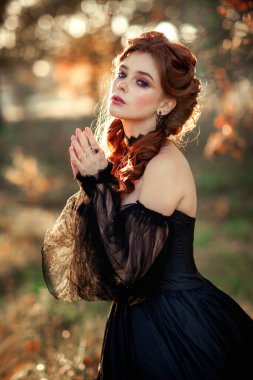 Sonbahar ağacının yanında duran muhteşem moda gotik kız portresi. Fantezi sanat çalışmaları. Siyah elbiseli ve şapkalı kızıl saçlı manken kamera ve pozlara bakıyor. 