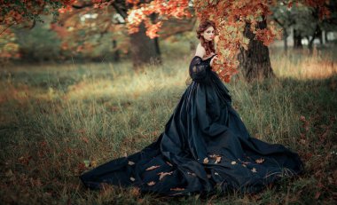 Sonbahar ağacının yanında duran muhteşem moda gotik kız portresi. Fantezi sanat çalışmaları. Siyah elbiseli ve şapkalı kızıl saçlı manken kamera ve pozlara bakıyor. 