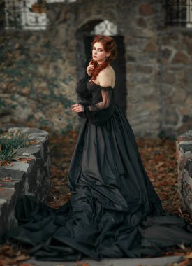 Taş duvarın yanında duran muhteşem moda gotik kız portresi. Fantezi sanat eserleri. Siyah elbiseli ve şapkalı kızıl saçlı manken kamera ve pozlara bakıyor. 