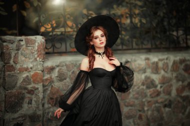 Taş duvarın yanında duran muhteşem moda gotik kız portresi. Fantezi sanat eserleri. Siyah elbiseli ve şapkalı kızıl saçlı manken kamera ve pozlara bakıyor. 