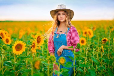 Güneş çiçekleri tarlasında poz veren genç ve güzel sarışın kız. Hasır şapkalı ve kot tulumlu güzel, şık, renkli kırsal resimlerde..