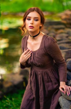 Beautiful red haired girl in brown long medieval dress and black vintage choker sitting on stone wall. Art work of romantic lady with book.Pretty tenderness model posing on nature. clipart