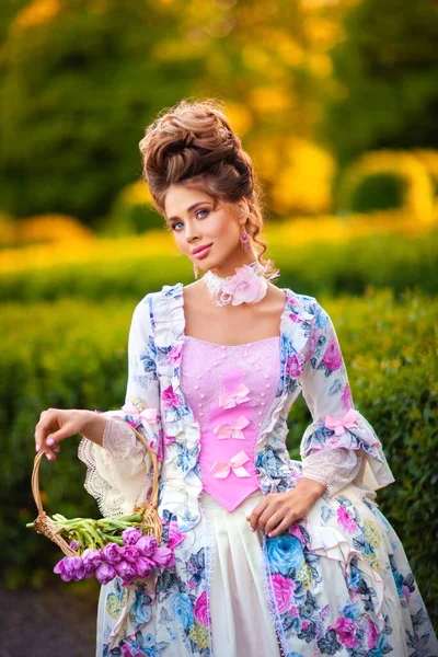 春花のバスケットと緑の日当たりの良い庭に立って完璧な髪型を持つ美しいロマンチックな女の子 妖精の美しいヴィンテージドレスのポーズで女王を夢見ます — ストック写真