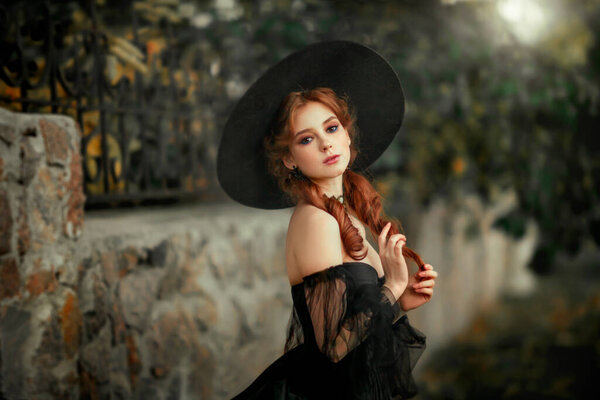 Portrait of magnificent Fashion gothic girl standing near stone wall .Fantasy art work.Amazing red haired model in black dress and hat looking at camera and posing.Fairytale about young princess