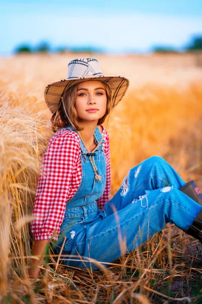 年轻漂亮的金发姑娘在田里摆姿势 戴着草帽 头戴牛仔布的漂亮的女牛仔在温暖多彩的乡村图画上穿工作服 — 图库照片