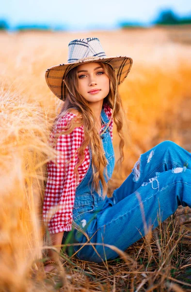 年轻漂亮的金发姑娘在田里摆姿势 戴着草帽 头戴牛仔布的漂亮的女牛仔在温暖多彩的乡村图画上穿工作服 — 图库照片