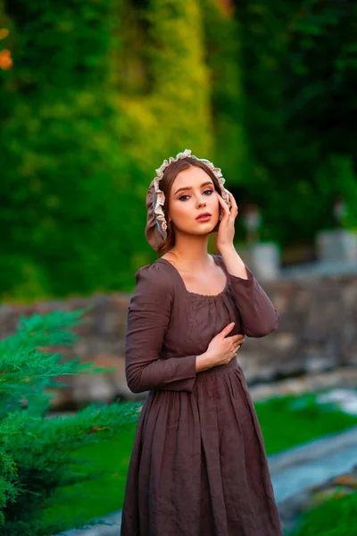 美丽的红头发姑娘穿着棕色的中世纪长裙 戴着帽子 站在石墙旁边 浪漫女士的艺术作品 以自然为特征的漂亮柔情模型 — 图库照片