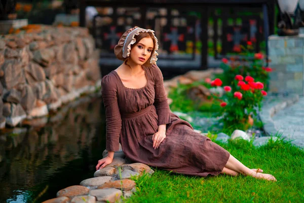 美しい赤髪の少女茶色の長い中世のドレスとボンネットの石の壁の近くに立っている ロマンチックな女性の芸術作品 自然の上にポーズかなり優しさモデル — ストック写真