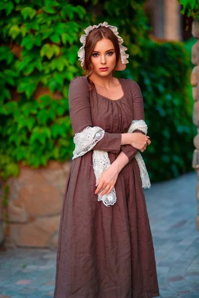 美丽的红头发姑娘穿着棕色的中世纪长裙 戴着帽子 站在石墙旁边 浪漫女士的艺术作品 以自然为特征的漂亮柔情模型 — 图库照片