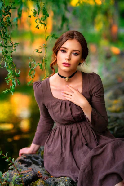 茶色の長い中世のドレスと石の壁に座って黒のヴィンテージチョーカーで美しい赤い髪の女の子 本を持つロマンチックな女性の芸術作品 かなり優しさモデルは 自然の上にポーズ — ストック写真