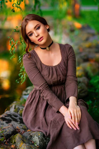 美丽的红头发女孩穿着棕色的中世纪长裙和黑色的古董车坐在石墙上 带着书的浪漫女子的艺术作品 以自然为写照的漂亮柔情模型 — 图库照片