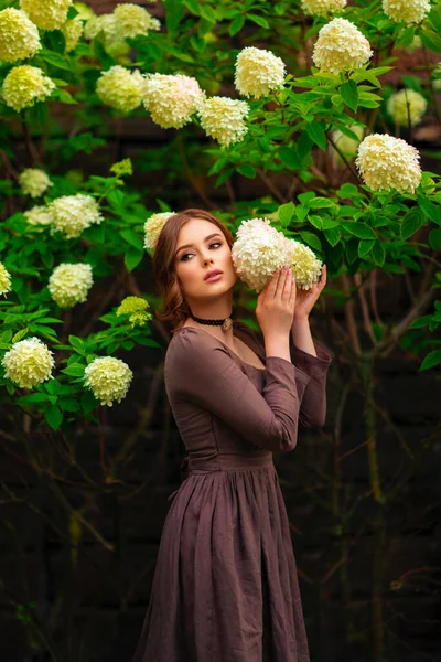 美丽的红头发姑娘穿着棕色的中世纪长裙 站在白花旁 浪漫女人的艺术作品 美丽的柔情模特儿在做梦 看着远方 — 图库照片