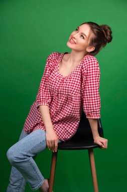 Yeşil arka planda sandalyede poz veren kareli gömlekli ve kot pantolonlu güzel bir genç kız izole edilmiş. Çekici bir modelle stüdyo çalışması..