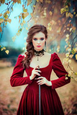 Sonbahar ormanlarında dikilen muhteşem moda gotik kız portresi. Fantezi sanatı. Kılıçlı kırmızı saçlı manken.. 