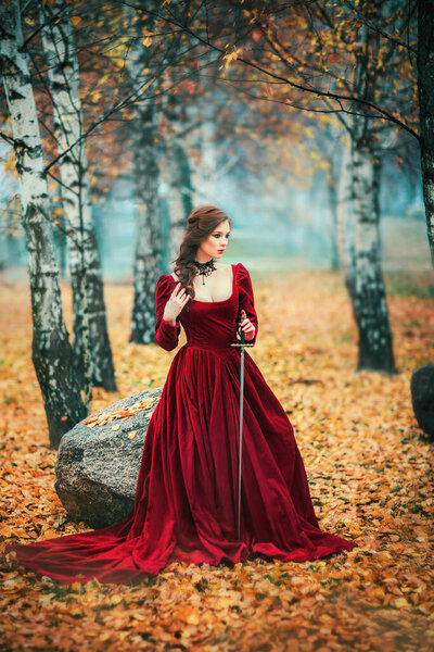 Portrait of magnificent Fashion gothic girl standing in autumn forest .Fantasy art work.Amazing red haired model in claret dress with a sword .Fairytale about young princess-warrior.