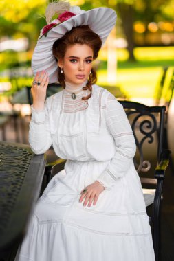 Güzel, kızıl saçlı, beyaz, uzun, klasik elbiseli ve şapkalı bir kız güneşli arka planda eski bir masanın yanında oturuyor. Mükemmel saç stili ve makyajı olan sevimli bir model. Sanat eserleri ve peri masalları