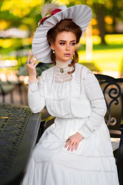 Güzel, kızıl saçlı, beyaz, uzun, klasik elbiseli ve şapkalı bir kız güneşli arka planda eski bir masanın yanında oturuyor. Mükemmel saç stili ve makyajı olan sevimli bir model. Sanat eserleri ve peri masalları