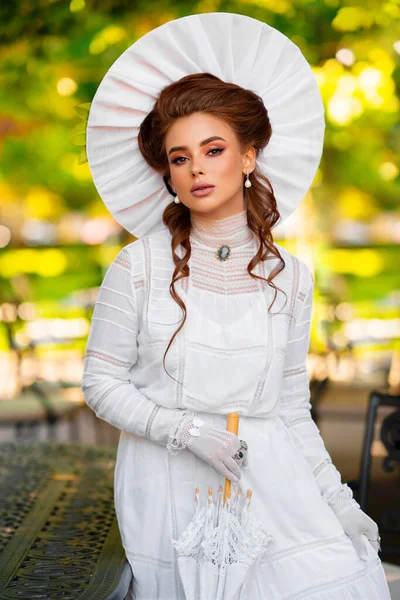 흰색긴 빈티지 드레스와 모자를 아름다운 소녀가 양지바른 배경에 빈티지 테이블 — 스톡 사진