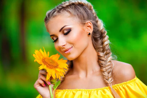 自然を楽しむ黄色のドレスの美しい若いブロンドの女の子 幸せな笑顔の女性がひまわりと緑の森の中に立っている 暖かい写真 美術作品 — ストック写真