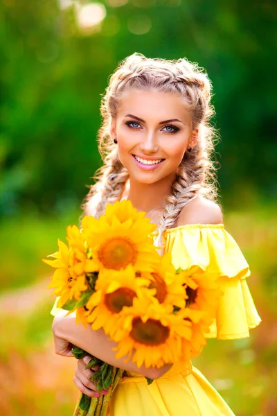自然を楽しむ黄色のドレスの美しい若いブロンドの女の子 幸せな笑顔の女性がひまわりと緑の森の中に立っている 暖かい写真 美術作品 — ストック写真