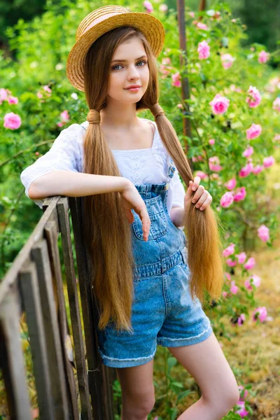 美丽的金发女郎穿着斜纹棉布工作服 戴着草帽 在玫瑰园里享受大自然的美景 年轻漂亮的模特站在篱笆边 背景上长着绿树和粉红的玫瑰 — 图库照片