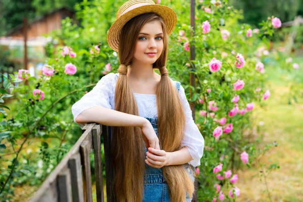ローズガーデンで自然を楽しむデニム全体と藁帽子を身に着けている美しい編組ブロンドの女の子 緑の木とピンクのバラを背景にフェンスの近くにポーズ若いかなりモデル — ストック写真