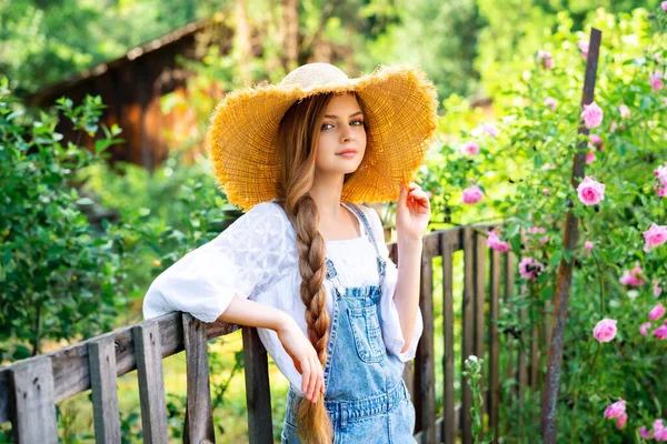 ローズガーデンで自然を楽しむデニム全体と藁帽子を身に着けている美しい編組ブロンドの女の子 緑の木とピンクのバラを背景にフェンスの近くにポーズ若いかなりモデル — ストック写真