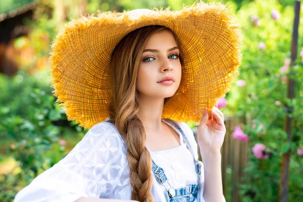 美丽的金发女郎穿着斜纹棉布工作服 戴着草帽 在玫瑰园里享受大自然的美景 年轻漂亮的模特 蓝眼睛 靠着篱笆站着 背景上有绿树和粉红的玫瑰 — 图库照片