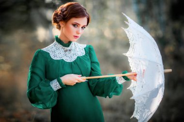 Çitin yanında duran yeşil, eski püskü elbiseli ve çiçek sepetli güzel kızıl saçlı kız. Mükemmel saç stilli, rüya gören ve kameraya bakan sevimli model. Sanat eseri ve peri masalı.