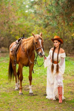 Ekstra uzun örgülü, dantel elbiseli, turuncu şapkalı ve at ile çiftlikte poz veren güzel bir kovboy kız. Kırsalda güzel bir modelin portresi. Sıcak sanat..