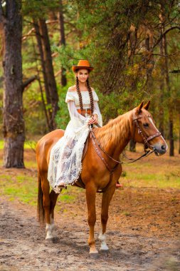 Ekstra uzun örgülü, dantel elbiseli, turuncu şapkalı ve at ile çiftlikte poz veren güzel bir kovboy kız. Kırsalda güzel bir modelin portresi. Sıcak sanat..