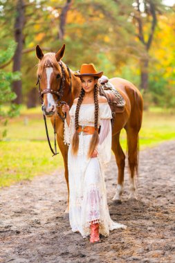 Ekstra uzun örgülü, dantel elbiseli, turuncu şapkalı ve at ile çiftlikte poz veren güzel bir kovboy kız. Kırsalda güzel bir modelin portresi. Sıcak sanat..