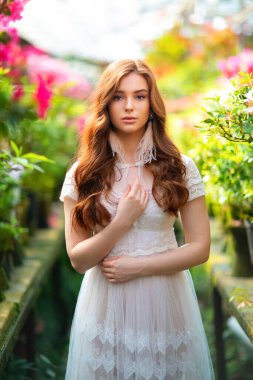 Renkli çiçeklerle dolu bir bahçede duran beyaz dantel elbiseli güzel kızıl saçlı kız. Romantik bir kadının sanat eseri. Kameraya bakan oldukça hassas bir model..