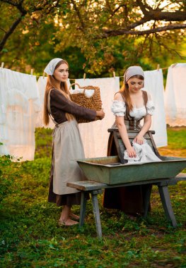  Ortaçağ kostümü giymiş iki genç çamaşırcı köyde eski bir yalakta elbiselerini yıkıyor. Kırsalda çalışan güzel kızlar. Peri masalı eserleri.