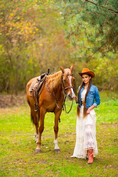 美丽的女牛仔 留着特别长的辫子头发 穿着老式花边衣服 头戴橙色的帽子 骑着马站在牧场上 一个漂亮的乡村模特儿的画像 温暖的艺术 — 图库照片